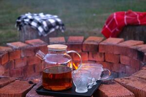 Transparent teapot with tea, cups on a tray on a brick fire pit in the courtyard of the house in a cozy summer evening. photo