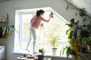 Woman manually washes the window of the house with rag cleaner and mop inside the interior with home plants on windowsill with attic. Restoring order and cleanliness in the spring, cleaning servise photo