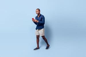 full body black young man look at smartphone go walking over blue background. photo