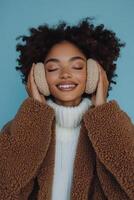 Young woman smiles while wearing warm ear muffs and a cozy sweater against a blue backdrop photo