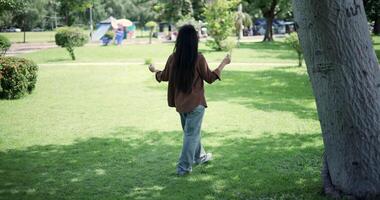 Handheld back view shot, Happy young woman enjoys a leisurely stroll, immersing herself in the nature around her in a park, enjoying the sunlight filtering through the trees along the way. video