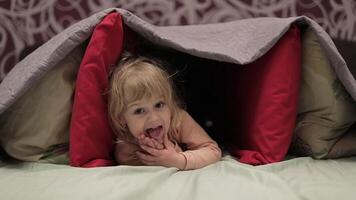 Little girl hides under blanket and pillows on the bed. Concept of hiding place video