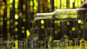 Barman pour frozen vodka from bottle into shot glass. Ice cubes against shiny gold party background video