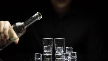 Barman pour frozen vodka from bottle into two glasses with ice. Black background video