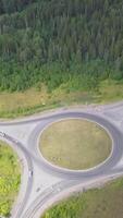 Top view of rural ring road with traffic. Clip. Scenic views of annular intersection with diverging roads in forest. Horizon with blue lake and coastal town. Travel and rest in summer video