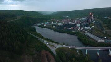 Top view of small factory town by river. Clip. Beautiful landscape with highway and river near town with factory in wooded area. Factory town by river with bridge in hilly forest area in summer video