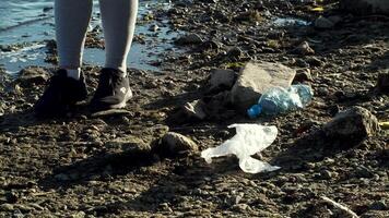 Garbage collection. Motion. A young woman cleaning up garbage on the beach with a river and bottles lying around. video