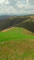 Top view of hilly mountains with green grass in summer. Shot. Beautiful green mountain slopes with grass and sparse forest on horizon. Landscape of mountains and hills on cloudy summer day video