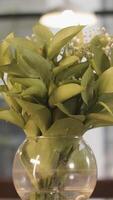 Gentle lilly of valley flowers in glass vase standing on wooden table on blurred background with windows and lamps. Stock footage. Spring may lily bouquet, decoration of room. video