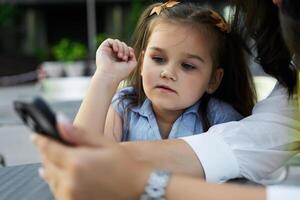 Young girl engrossed in a smartphone while sitting at a table with an adult during a sunny afternoon photo