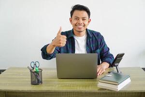 Happy smiling young asian man sitting and working on a laptop showing thumb up sign isolated on white background photo