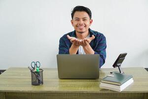 smiling young asian businessman sitting at workplace open both hands as if holding something photo
