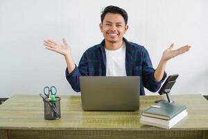 young asian businessman sitting at workplace having doubts with both arms open to the sides photo