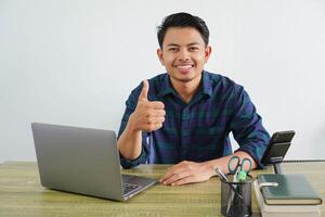 Smiling young asian man in blue shirt sit work at wooden desk with pc laptop isolated on white background. Achievement business career lifestyle concept. showing thumb up photo