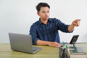 puzzled young asian man sit work at wooden desk with pc laptop. Achievement business career lifestyle concept. pointing index finger aside photo