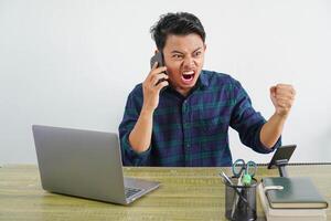 Angry irritated young asian man sit work at wooden desk with pc laptop. Achievement business career lifestyle concept. talking on mobile phone photo