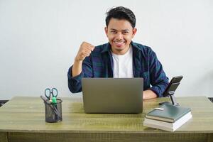 happy young asian businessman sitting at workplace an expression of enthusiasm working with his hands clenched into fists photo