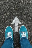 Feet in turquoise shoes standing on asphalt by a white directional arrow pointing forward photo