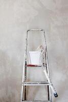 A white paint bucket with a used roller, positioned on a spattered paint step ladder against the concrete unpainted wall. Home renovation DIY essential tools. Minimalistic concept. Copy space. photo