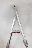 Detail of spattered paint step ladder with red step staying against the concrete unpainted wall. Home renovation DIY essential tools. Minimalistic concept. Copy space. photo