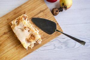 Piece of traditional thanksgiving apple cinnamon pie on the wooden cutting board on the white table, surrounded by fresh apples and walnuts. Autumn dessert. Closeup. Copy space. Top view photo