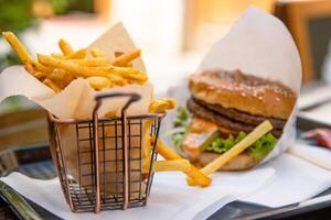 Closeup of a restaurant table with classic fast food meal. Burger with beef patty, fresh lettuce and a sesame seed bun wrapped in paper. Crispy golden French fries served on a tray. Selective focus photo