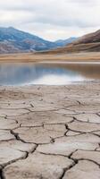 Dried cracked earth near a serene lake bordered by mountains on a cloudy day photo
