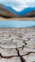 Dried cracked earth near a serene lake bordered by mountains on a cloudy day photo