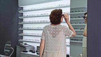 Sunglasses selection explored by woman, checking reflection, surrounded by different designs, browsing for style choice, modern interior, eyewear shopping experience, deciding on look video