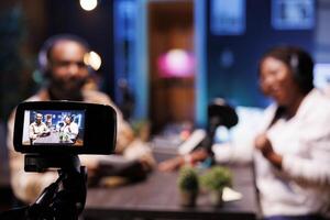 During live talk show broadcast on streaming platforms, cohosts use a professional camera for recording. African vloggers seen in blurry background using modern recording equipment to make a vlog. photo