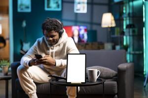 Focus on mockup tablet with man in blurry background sending messages on phone and listening music. Close up of isolated screen device in front of person wearing headphones, texting friends photo
