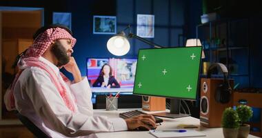 Arabic man looking at chroma key computer screen while working from home. Self employed person typing on keyboard, sending emails on green screen device in apartment office with TV as background noise photo