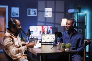 Black man with notebook is interviewing social media star and using audio equipment to record the program. Online radio host casually talking with influencer in home studio, recording podcast episode. photo
