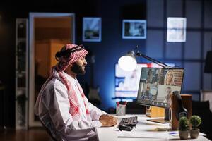 Image shows a Middle Eastern guy dressed in traditional attire, engrossed in an online meeting with his coworkers. Side-view shot of a youthful Arab man participating in a conference call. photo