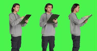 Asian guy reads information on files, thinking about annotations on clipboard papers or taking notes for completion. Young man writing on forms, standing against greenscreen backdrop. photo