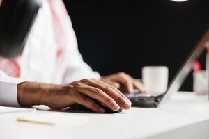Close-up view of a pair of male hands using a wireless mouse and laptop, doing online research. Detailed shot of a person with Arabian clothing making use of the minicomputer on the table. photo