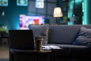 Focus on laptop and mini house plant on coffee table with television screen in blurry background. Neon lit apartment with cozy couch in front of smart TV displaying news, close up shot photo