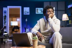 BIPOC remote worker sitting at home on couch late at night, overthinking about problems. Person struggling to finish work project before deadline, trying to come up with solutions photo