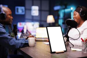 Male and female content creators wearing headphones are live streaming, while tablet displays blank screen. Black podcasters using equipment on desk while device shows copyspace mockup template. photo