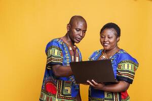 Smiling african american couple wearing ethnic clothes holding laptop and browsing internet together. Cheerful man and woman using portable computer and reading message online photo