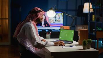 Arab remote worker inputting data on mockup laptop while working from home. Self employed freelancer performing tasks on isolated screen device in apartment office with TV as background noise photo