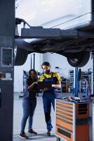 Repairman in car service orders new wheels for damaged vehicle using laptop. Engineer next to customer looking online for components to replace old ones in malfunctioning automobile on overhead lift photo