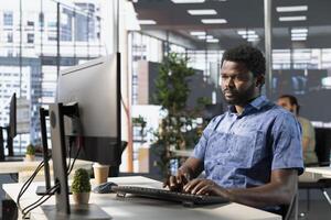 Startup employee sitting at desk in office, doing research for project with deadline approaching. African american man looking at computer in workspace, solving tasks during job shift photo