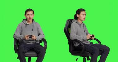 Asian guy using controller for gameplay, sitting on chair against greenscreen backdrop in studio. Young man enjoying online gaming plan with friends, gamer playing rpg games. photo