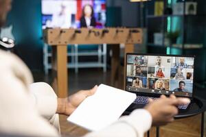 Management executive analyzing paperwork while holding videoconference meeting with remotely working multiethnic team. Manager teleworking in apartment, discussing project progress with workers photo