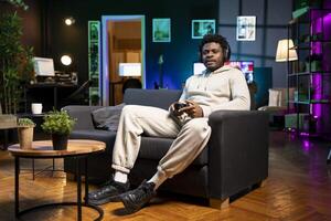 BIPOC man in dimly lit apartment playing videogames, relaxing and having fun. Gamer battling enemies in multiplayer game on gaming console during leisure time at home photo