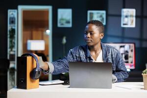 Young black woman working from home using a laptop and placing a sticky note on a speaker. African American blogger putting post-it notes on a device while using her personal computer. photo