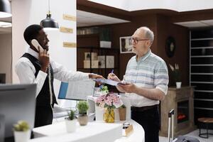 Elderly retired caucasian guy signs reservation paperwork at counter of hotel lobby while african american employee takes a call. Senior customer fills out check-in papers as receptionist is on phone. photo