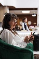 Detailed view of elderly female client seated in hotel foyer, using her cell phone to browse the internet. Retired old woman checking in online using mobile device. Close-up, side-view. photo