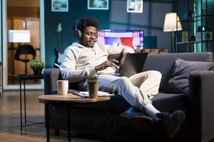 Happy remote worker listening to music on headphones while working from home. Relaxed smiling freelancer enjoying himself, typing on computer in stylish apartment personal office photo